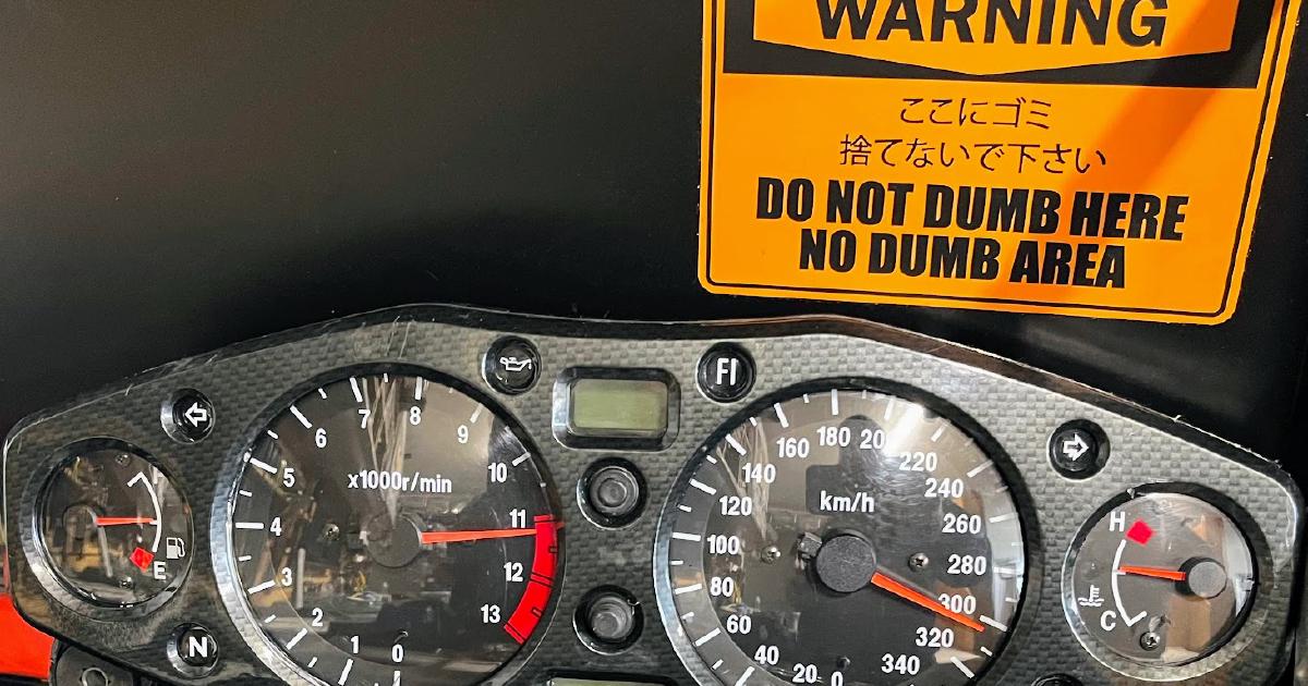 Motorcycle gauge cluster mounted on a garage wall, speedometer needle near 300 km/h, beside a warning sticker reading 'Do not dumb here, no dumb area'.
