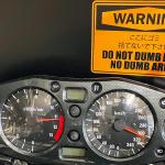 Motorcycle gauge cluster mounted on a garage wall, speedometer needle near 300 km/h, beside a warning sticker reading 'Do not dumb here, no dumb area'.