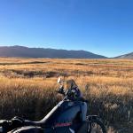 Adventure motorbike parked beside a golden tussock plain at sunrise, low sun flaring over mountain ranges on the horizon under a clear blue sky.
