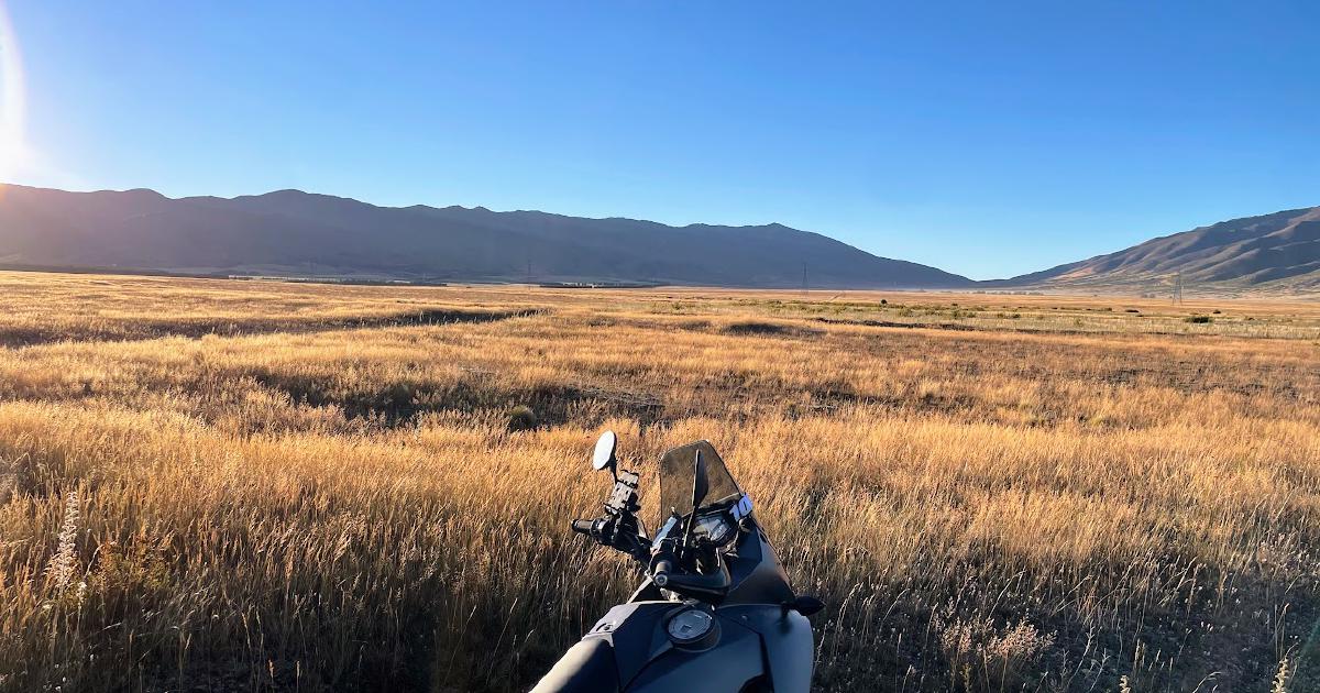 Adventure motorbike parked beside a golden tussock plain at sunrise, low sun flaring over mountain ranges on the horizon under a clear blue sky.