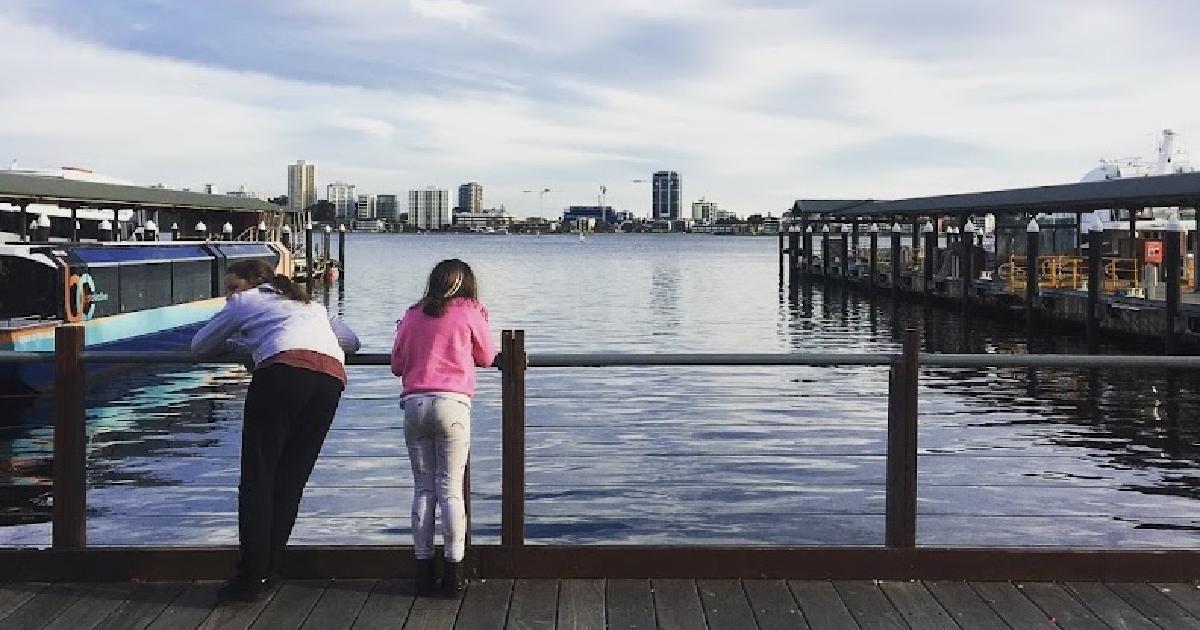 Two kids lean on a wharf railing, looking out across the water at a distant city skyline.