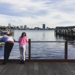 Two kids lean on a wharf railing, looking out across the water at a distant city skyline.