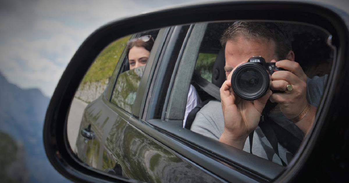 A self-portrait reflected in a car's wing mirror - me raising a DSLR to photograph my own reflection, my partner in the passenger seat behind, with mountains and a cloudy sky in the background.