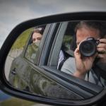 A self-portrait reflected in a car's wing mirror - me raising a DSLR to photograph my own reflection, my partner in the passenger seat behind, with mountains and a cloudy sky in the background.
